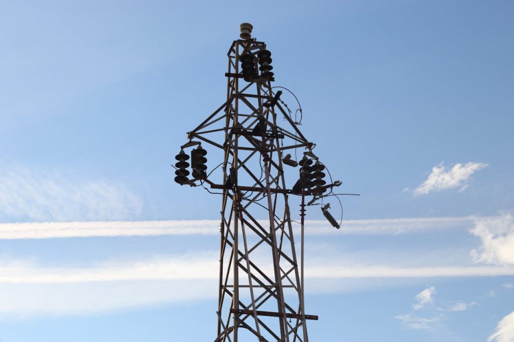 A high voltage power line tower set against a clear blue sky with visible clouds.
