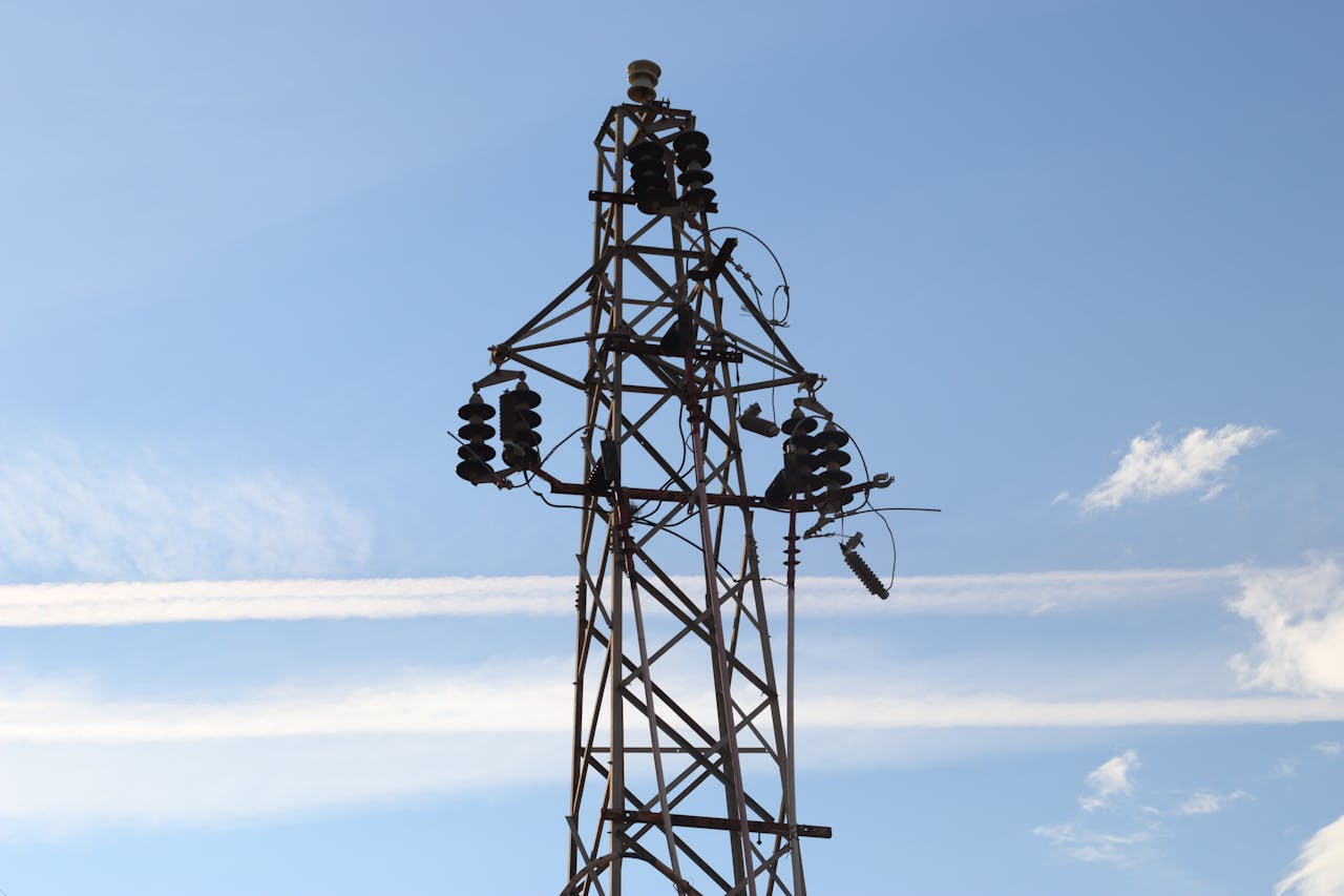 A high voltage power line tower set against a clear blue sky with visible clouds.